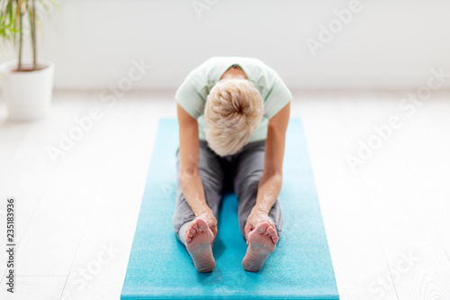 Mature woman in joga pose exercise in appartment at rug with window on background