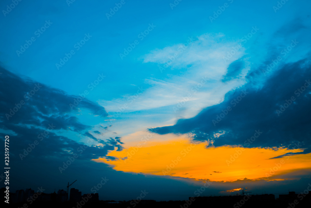 Atmospheric blue cloudy sky behind silhouettes of city buildings. Cobalt and orange background of sunrise with dense clouds and bright yellow sunny light for copy space. Cyan heaven above clouds. Atmospheric blue cloudy sky behind silhouettes of city buildings. Cobalt and orange background of sunrise with dense clouds and bright yellow sunny light for copy space. Cyan heaven above clouds.