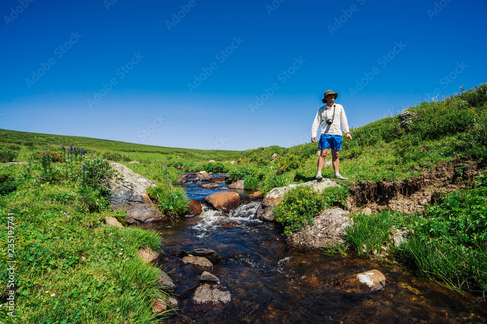 Traveler with camera on stone in mountain creek. Adventure of tourist. Hiking in mountains. Rich vegetation of highlands. Stream of clear water in brook. Vivid sunny landscape of majestic nature.