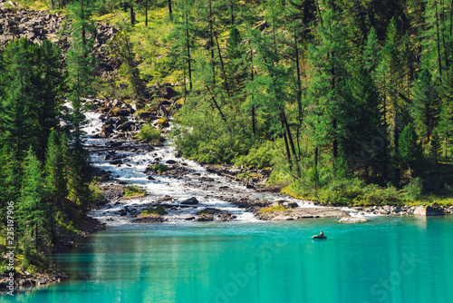 Fototapeta Naklejka Na Ścianę i Meble -  Mountain creek flows into lake. View above fisherman in boat on azure water. Wonderful forest in sunlight. Big boulders in brook. Atmospheric beautiful landscape of highland nature in sunny day.