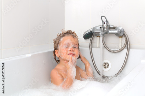 Little blonde girl in the bathroom with foam on her face. Eyes are closed. Independent baby taking a bath on white background. Infant washing and bathing. Hygiene and health care.