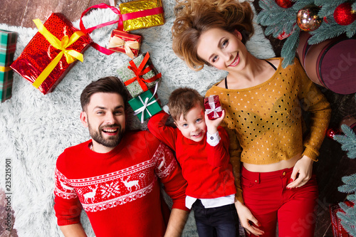 Happy family: wife woman female, husband man male in warm sweater and son boy lying on carpet floor with christmas presents and enjoying celebrating winter holidays: Christmas Eve and Happy New Year.