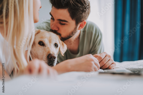 Pleasant bearded man kissing his dog while lying in bed with his girlfriend