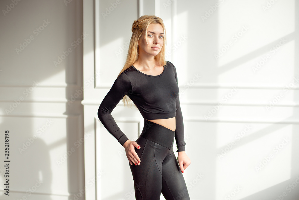 Young slim woman with an athletic body long blonde hair wearing in black  sports sportswear top and leggings standing in bright yoga room with big  panaramic window preparing before training health-life Stock