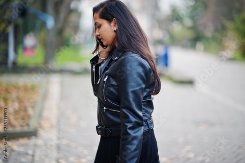 Pretty indian girl in black saree dress and leather jacket posed outdoor at autumn street and speaking on mobile phone.