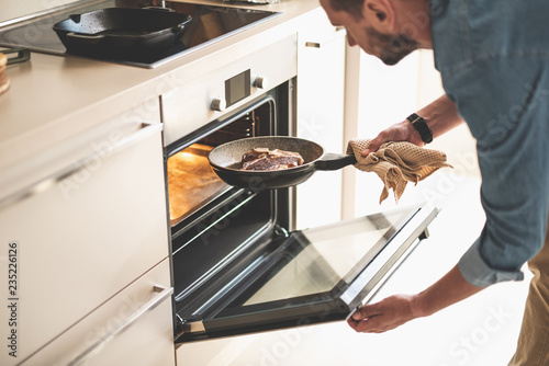 Gentleman holding frying pan with beef steak while opening oven door