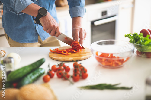 Fototapeta Naklejka Na Ścianę i Meble -  Cooking dinner at home. Close up of male hands slicing red bell pepper with knife on cutting board
