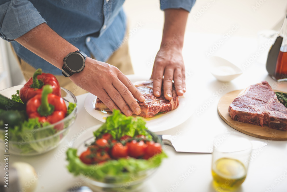 Close up of male hands greasing beef steak with olive oil. Fresh vegetables, knife and cutting board on white desk Close up of male hands greasing beef steak with olive oil. Fresh vegetables, knife and cutting board on white desk