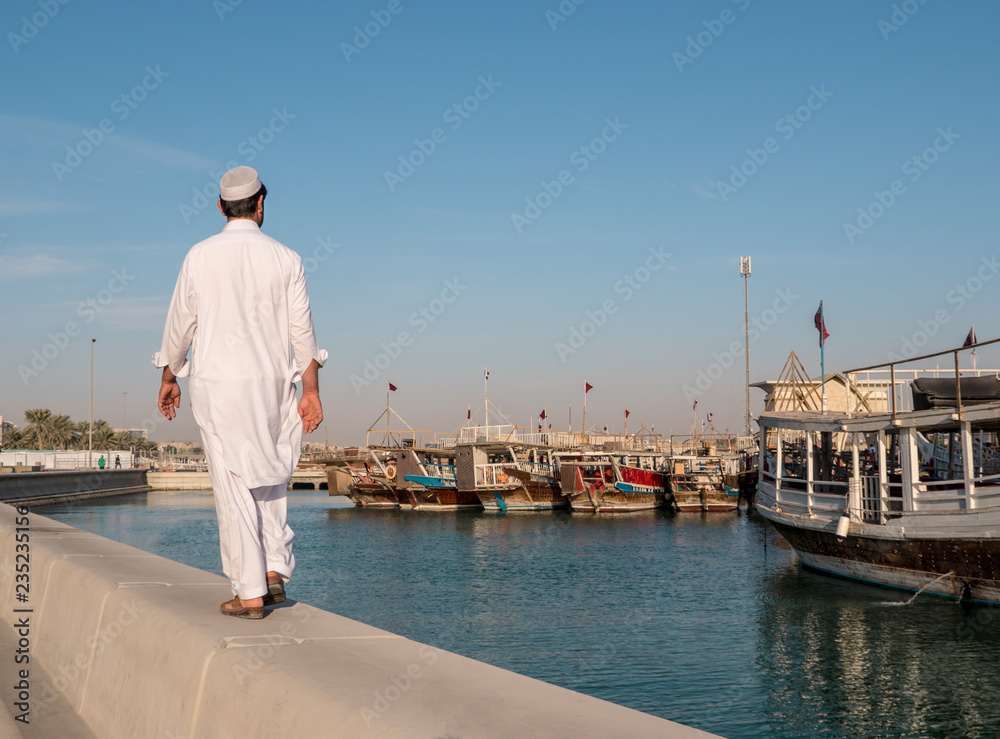 Middle eastern Man Walking by Corniche road in Doha. View with Traditional Wooden Boats with Floating Qatar Flags. Corniche Broadway. Qatar, Middle East