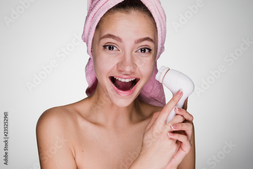 joyful laughing girl with a pink towel on her head holding an electric brush for deep cleansing of the face skin