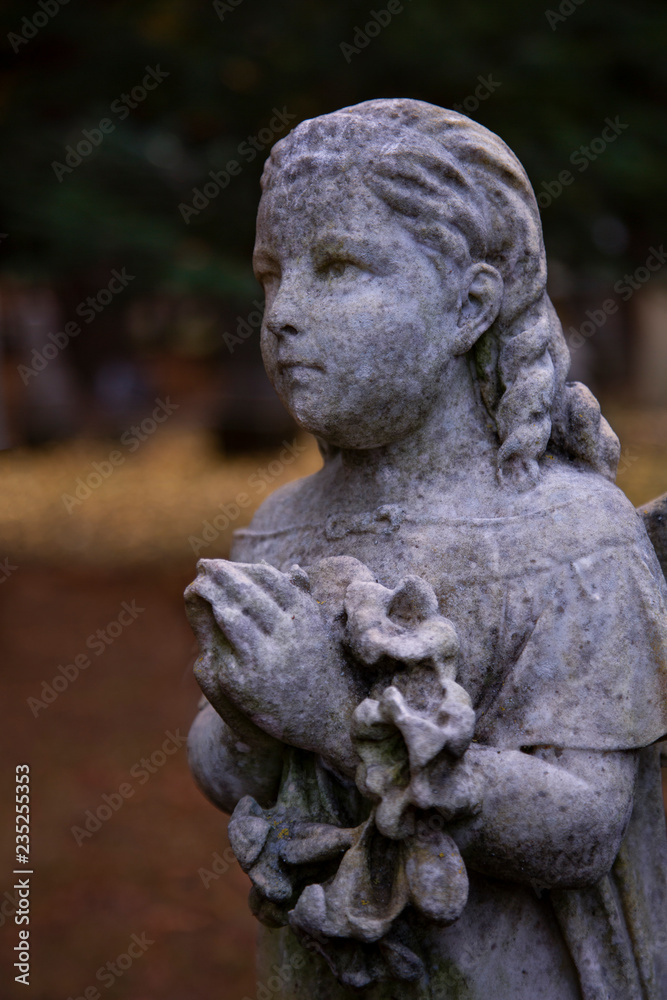Partial View of Victorian Statuary, a Girl's Face, Shoulders, and Hands in Prayer Position Holding a Wreath with Out of Focus Foliage and Trees in Background