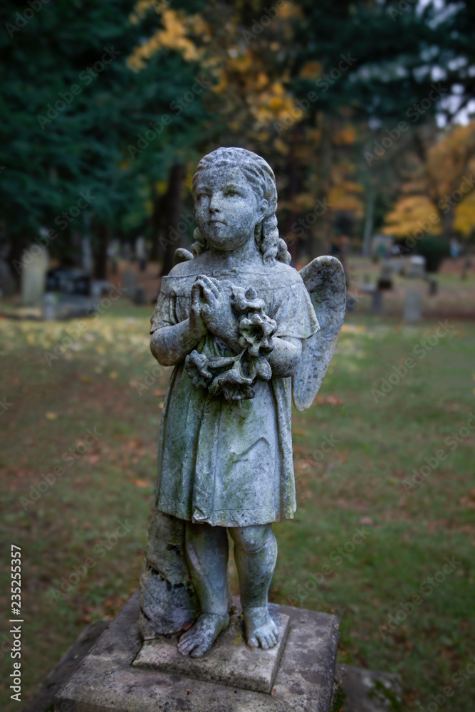 Full View of Victorian Statuary Covered in Green Moss, a Girl with Angels' Wings, Hands in Prayer Position Holding a Wreath with Out of Focus Cemetery in Background in Vertical Format