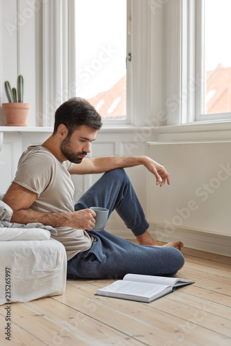 Shot of attractive clever man enjoys reading book at home sits at floor near bed, drinks fresh hot beverage, likes novel, feels inspired and realxed, enjoys calm atmosphere. Literature develops us