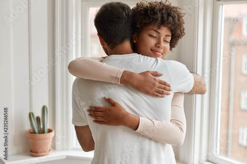Valokuva Pleased dark skinned young woman gives warm hug to her boyfriend, being pleased, pose near window, have romantic relationship, stand in cozy room