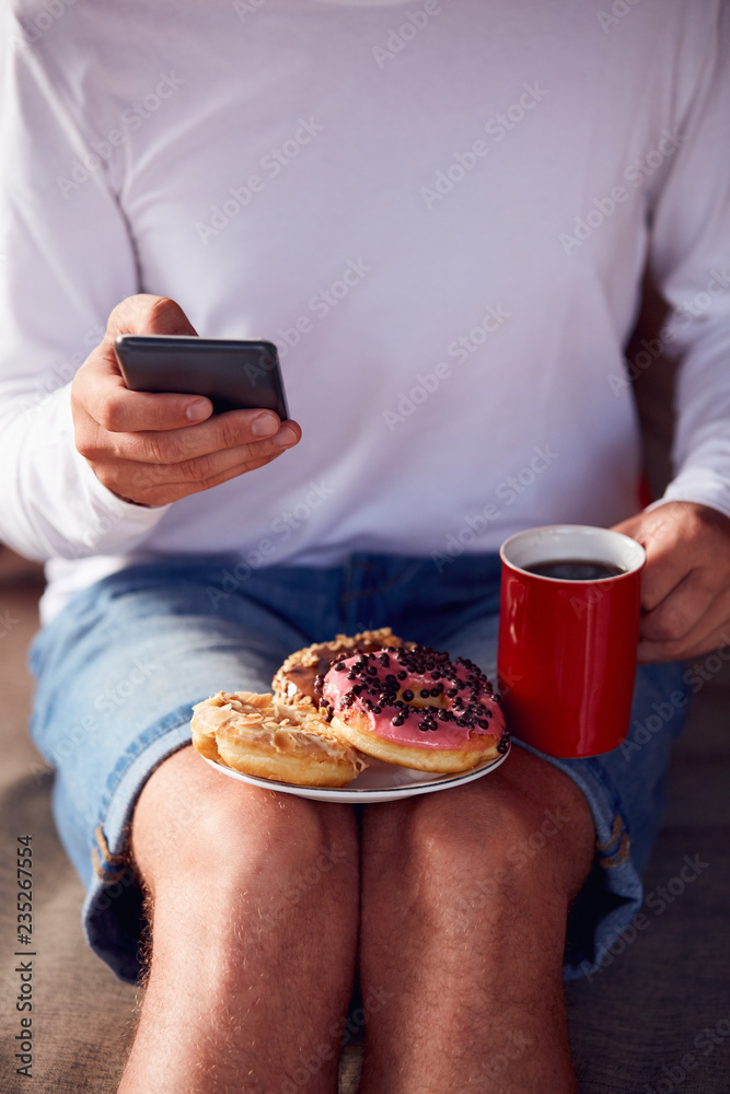 Man sitting on a terrace sofa and drinking coffee/tea, eating donuts while using cellphone.