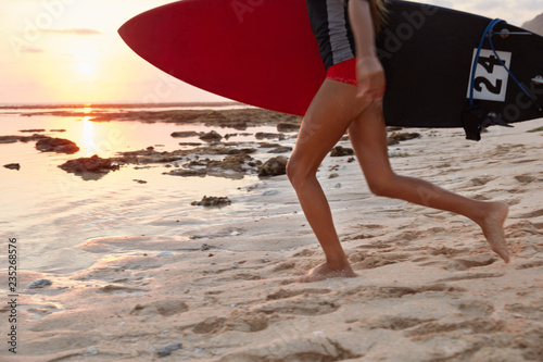 Photo of unrecognizable young woman likes surfing, runs on beach, carries surfboard under arm, wears swimsuit, beautiful sunset near ocean at evening. Extreme sport and surfing concept. Sea view