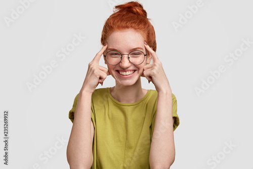 Use your brain. Happy red haired girl holds both index fingers on temples, tries to think before acting stupidly, smiles happily, dressed in casual summer clothes, stands against white background. photo