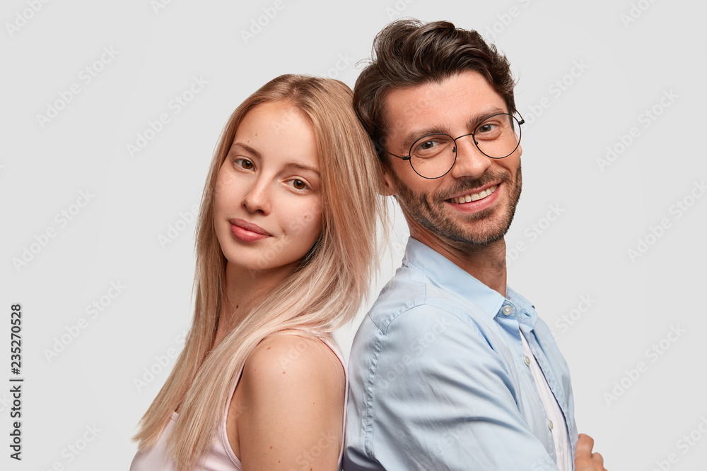 Lovely couple stand back to each other, being in good mood, isolated over white background. Pretty blonde lady, unshaven cheerful man in spectacles being partners, feel support, ready to help Lovely couple stand back to each other, being in good mood, isolated over white background. Pretty blonde lady, unshaven cheerful man in spectacles being partners, feel support, ready to help