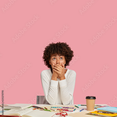 Vertical shot of shocked dark skinned professional multiplier covers mouth with both hands, wears white sweater, forgets about importnat task, sits at workplace copy space above for your advertisement photo