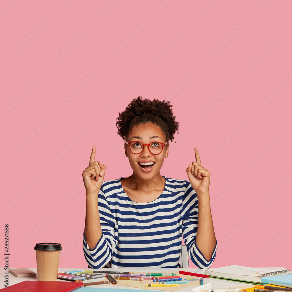 Vertical shot of glad dark skinned lovely woman artist, points with both index fingers, wears striped sweater, models against pink background, feels excited, poses at desktop with spiral notebook Vertical shot of glad dark skinned lovely woman artist, points with both index fingers, wears striped sweater, models against pink background, feels excited, poses at desktop with spiral notebook