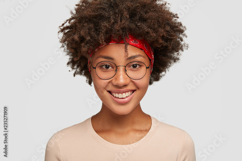 Close up shot of friendly looking cheerful African Amrican female with tender expression, pleasant smile, rejoices amazing trip on summer vacation, wears round glasses, models in white studio