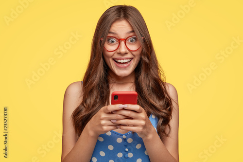 Shot of cheerful female model wears glasses, holds cell phone, takes pictures, talks via video message, has nice friendly talk, dressed in fashionable outfit, isolated over yellow background photo