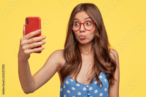 Pretty girl takes selfie, pouts lips at camera of cell phone, makes video call, flirts with boyfriend, shoots something for blog, takes photo of herself, wears glasses, models against yellow wall