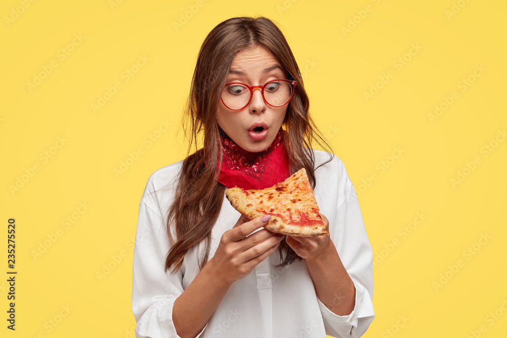Shocked emotive lady looks with surprisement at slice of pizza, amazed with its wonderful taste, wears spectalces and white shirt, models against yellow wall. People, reaction and nutrition concept