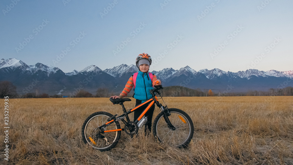 One caucasian children walk with bike in wheat field. Little girl walking black orange cycle on background of beautiful snowy mountains. Biker stand with backpack and helmet. Mountain bike hardtail.