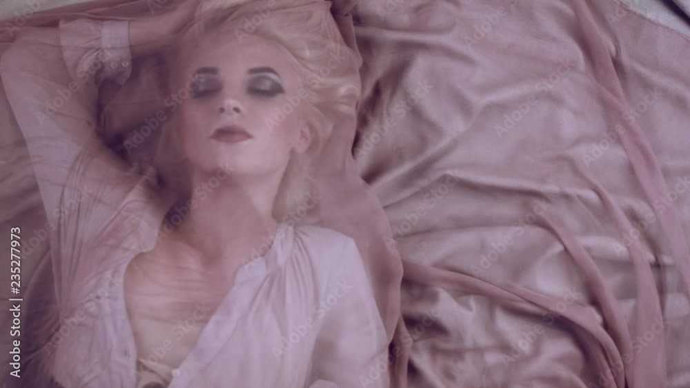 Beautiful young tender girl in chiffon dress lies under thin pink fabric that develops from the wind. Sensual image of girl with gentle make-up. Advertising spirits, textiles. Beautiful make-up.