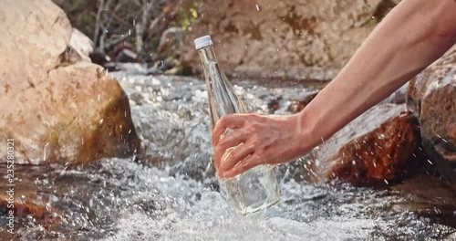 Human hand taking a glass bottle out of water stream of beautiful fresh river, flowing in mountains with murmuring and sunbeams - harmony with nature 4k photo