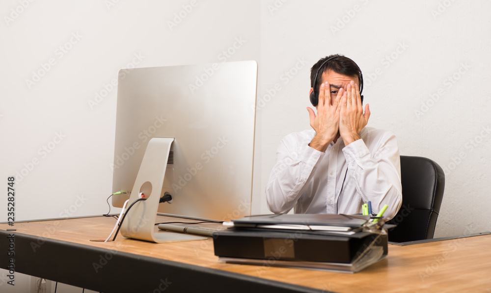 Telemarketer man in a office covering eyes by hands and looking through the fingers Telemarketer man in a office covering eyes by hands and looking through the fingers