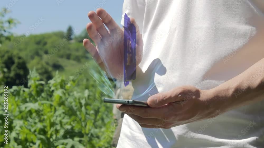 Man shows concept hologram adapt on his phone. Person in white t-shirt with future technology holographic screen and green nature background Man shows concept hologram adapt on his phone. Person in white t-shirt with future technology holographic screen and green nature background