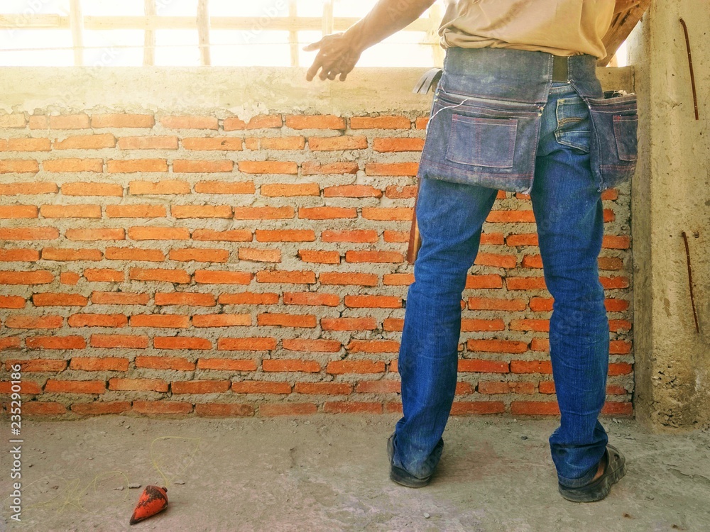 Part of back side mason man in brown t shirt, blue jeans and tool belt bag is working in construction site with flare light on surface of bricks wall, selective focus with copy space