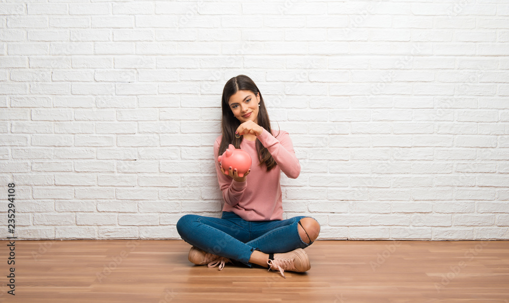 Teenager girl sitting on the floor in a room taking a piggy bank and happy because it is full Teenager girl sitting on the floor in a room taking a piggy bank and happy because it is full