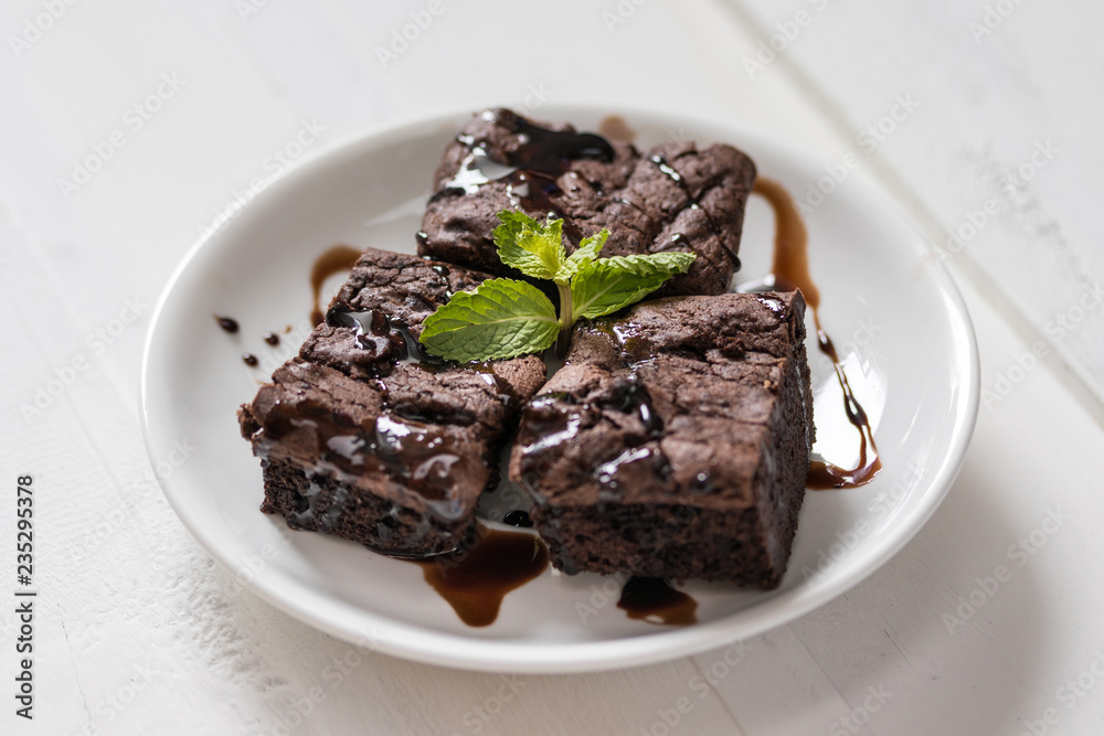 A plate of brownies with chocolate sauce on white wooden background, mint leaf on top,