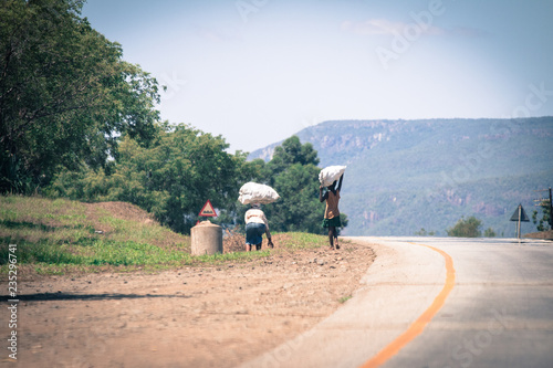 Zwei Menschen aus Afrika mit schweren Säcken auf dem Kopf laufen eine Straße entlang © marksn.media