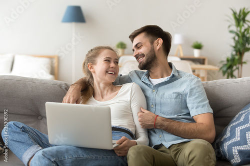 Happy millennial couple cuddle at cozy couch at home using laptop, smiling husband and wife relax on comfortable sofa spending weekend together, loving man and woman hug enjoying leisure time