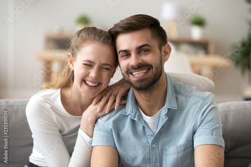 Portrait of smiling young husband and wife hugging sitting on couch, happy millennial couple look at camera posing for family picture at home, laughing man and woman hug having fun together
