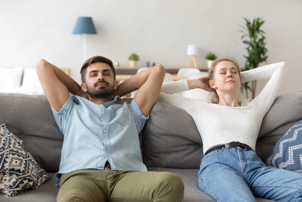 Calm husband and wife lying on cozy couch hands over head, peaceful millennial couple stretch relaxing on comfortable sofa with eyes closed, man and woman rest in living room taking nap or dreaming