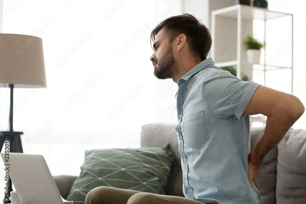 Tired millennial man sit on couch having back pain massaging to relieve symptoms, young male stretch suffering from spinal spasm or strain working too long in incorrect posture. Sedentary life concept Tired millennial man sit on couch having back pain massaging to relieve symptoms, young male stretch suffering from spinal spasm or strain working too long in incorrect posture. Sedentary life concept