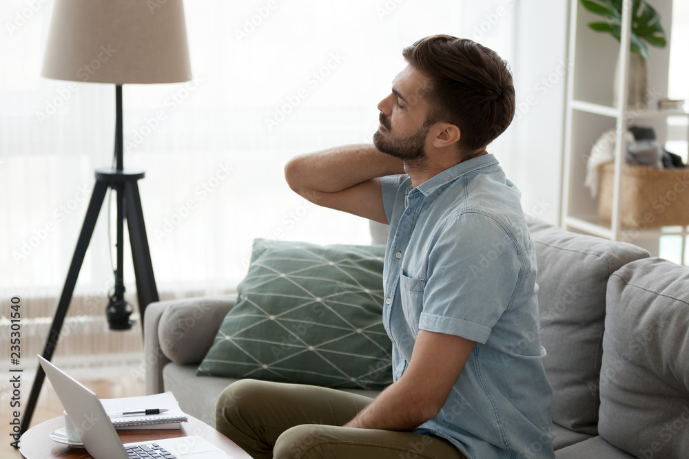 Tired male sit on couch massaging neck suffering from pain or strain  sitting in incorrect posture, man using laptop working long hours on sofa  having back spasm symptoms. Sedentary lifestyle concept Stock