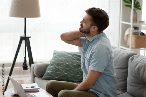 Tired male sit on couch massaging neck suffering from pain or strain sitting in incorrect posture, man using laptop working long hours on sofa having back spasm symptoms. Sedentary lifestyle concept photo