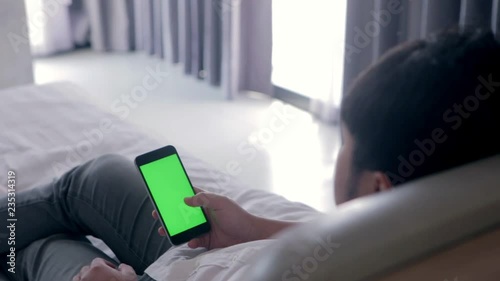 Back view of young asian handsome man using smartphone with green screen laying down on bed while chatting with friend