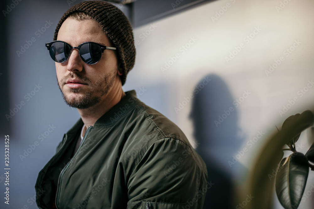 Young Serious Trendy Guy Sitting in Lobby Wearing a Green Jacket, Sunglasses, and a Beanie Young Serious Trendy Guy Sitting in Lobby Wearing a Green Jacket, Sunglasses, and a Beanie