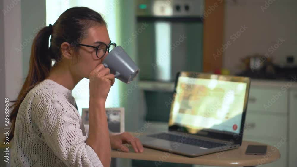 Girl sits on kitchen freelancing working at home searching information on laptop and drink coffee Girl sits on kitchen freelancing working at home searching information on laptop and drink coffee