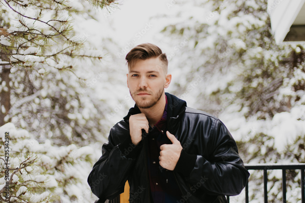 Portrait of Attractive Trendy Modern Young Male Model Smiling and Enjoying the Outdoors and the White Winter Snow in the Tree Canopy While Posing in Fashionable Leather Bomber Jacket Portrait of Attractive Trendy Modern Young Male Model Smiling and Enjoying the Outdoors and the White Winter Snow in the Tree Canopy While Posing in Fashionable Leather Bomber Jacket