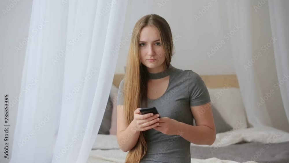 Joyful young beautiful woman, girl in the bedroom sitting on the bed near the window and looks out the window and listening music on her smartphone. Joyful young beautiful woman, girl in the bedroom sitting on the bed near the window and looks out the window and listening music on her smartphone.