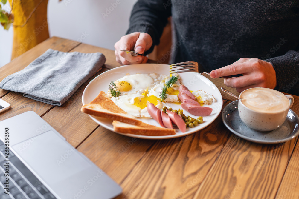 The man eat. Knife and fork in hand. The concept of eating at work. Laptop and Breakfast on the table.. American style breakfast with fried eggs, sausage, green peas and toast.