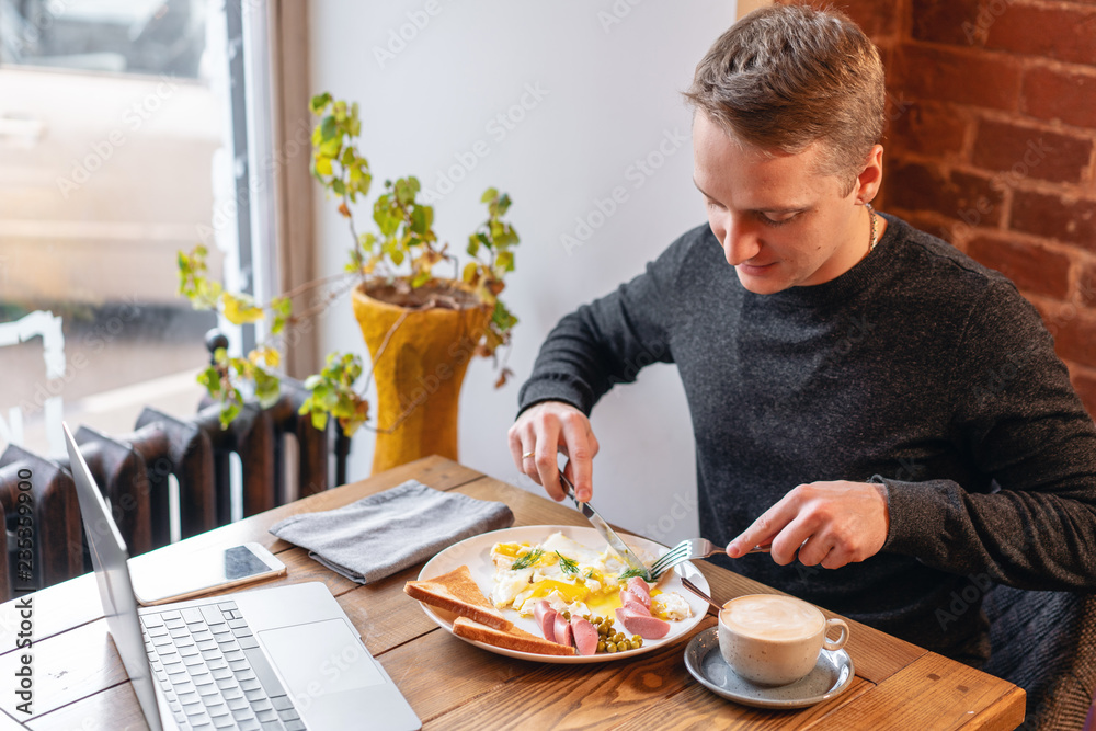 The man eat. Knife and fork in hand. The concept of eating at work. Laptop and Breakfast on the table.. American style breakfast with fried eggs, sausage, green peas and toast. The man eat. Knife and fork in hand. The concept of eating at work. Laptop and Breakfast on the table.. American style breakfast with fried eggs, sausage, green peas and toast.